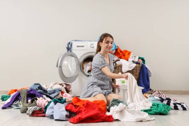 Gri arka planda çamaşır makinesinin yanında kirli çamaşırları sıralayan genç bir kadın.