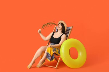 Palmiye yaprağı ve şişirilebilir yüzükle turuncu arka planda güverte koltuğunda oturan güzel genç kadın.