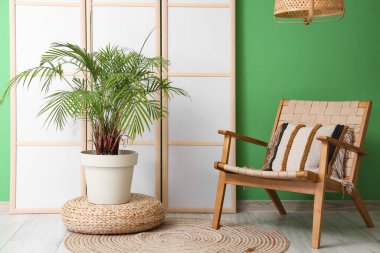 Yeşil duvarın yanında katlanır ekran, sandalye ve çiçek bulunan odanın içi.