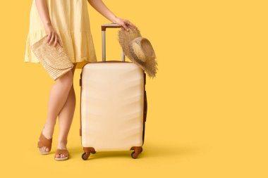 Sarı arka planda bavulu ve hasır şapkası olan genç bir kadın.