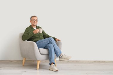 Beyaz duvarın yanındaki koltukta oturan bir fincan çayla yaşlı adam.