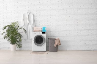 Modern çamaşır makinesi, çamaşır sepeti, ev bitkisi ve bornoz.