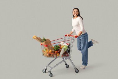 Gri arka planda alışveriş arabası olan güzel, genç ve mutlu bir kadın.