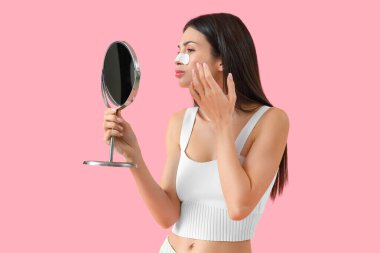 Burun bandı takmış genç bir kadın pembe arka planda aynaya bakıyor.