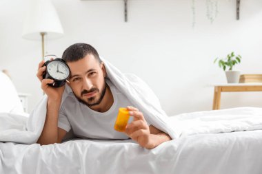 Çalar saati ve hapları olan stresli genç bir adam yatak odasında yatıyor.