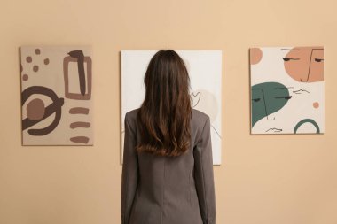 Sanat galerisindeki bej duvardaki tabloların yanında genç bir kadın.