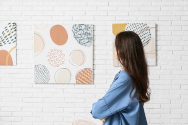 Sanat galerisindeki beyaz tuğla duvardaki resimlere bakan genç bir kadın.