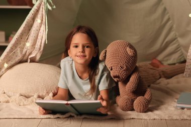 Kitabı ve oyuncak ayısı olan sevimli küçük bir kız evdeki çadırda yatıyor.