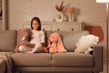 Oturma odasında oyuncaklarla kanepede oturmuş kitap okuyan tatlı küçük kız.