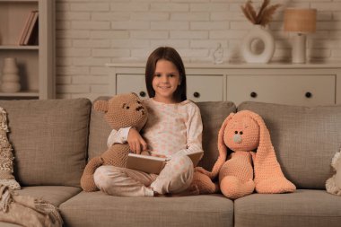 Oturma odasında oyuncaklarla kanepede oturmuş kitap okuyan tatlı küçük kız.