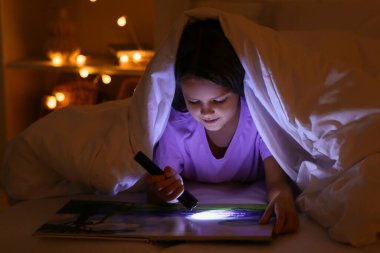 Evde el feneriyle yatakta masal okuyan tatlı küçük bir kız.