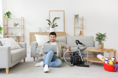 Evde dizüstü bilgisayarı ve elektrikli süpürgesi olan genç bir adam.