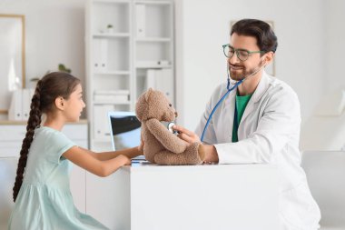 Steteskoplu erkek çocuk doktoru. Klinikte oyuncak ayı ve tatlı kızı dinliyor.