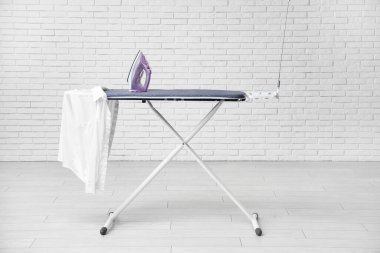 Beyaz tuğla duvarın yanında elektrikli ütü ve gömlekli bir tahta.