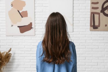 Sanat galerisinde beyaz tuğlalı duvardaki resimlere bakan genç bir kadın.