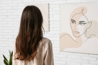 Sanat galerisi sergisinde, tabloların yanında beyaz tuğlalı duvara asılı genç bir kadın.