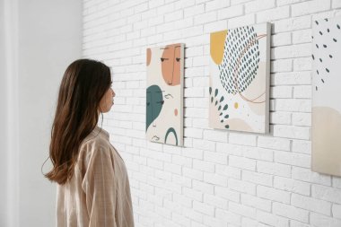 Sanat galerisindeki beyaz tuğla duvardaki resimlere bakan genç bir kadın.