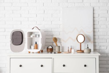 Cilt ürünleri olan küçük bir kozmetik buzdolabı, odadaki beyaz tuğla duvarın yanındaki çekmecelerin üzerindeki ayna ve resim. Yakın çekim.
