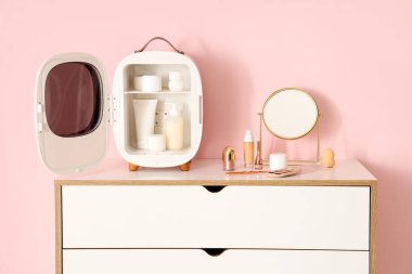 Küçük bir kozmetik buzdolabı aç. Masada pembe arka planda ürünler ve aynalar olsun. Yakın çekim.