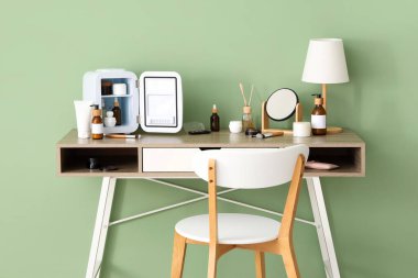 Küçük kozmetik buzdolabı, ayna ve lamba olan masa yeşil duvarın yanında.