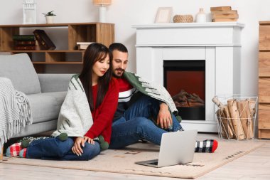 Evlerinde şöminenin yanında oturan ekose ve dizüstü bilgisayarlı mutlu çifti seviyorlar.