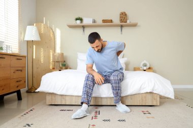 Evde sırt ağrısı çeken pijamalı genç bir adam var.