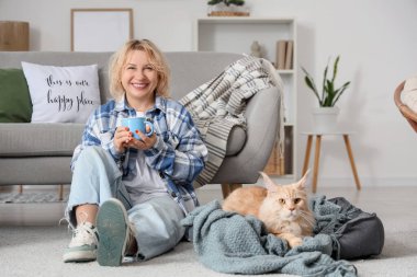 Güzel orta yaşlı bir kadın, sevimli bir kedi ve bir fincan çayla evde oturuyor.