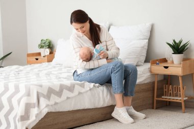 Genç anne yatak odasında beşikteki şapkayla küçük bebeğinin saçını tarıyor.