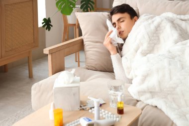 Evdeki kanepede hapşıran ekose ve peçeteli hasta genç adam.