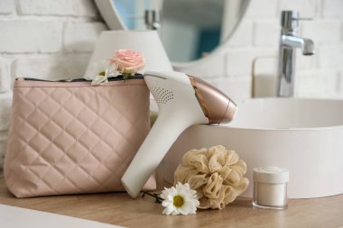 Kozmetik çantalı modern fotoepilatör, banyo süngeri ve banyo lavabosunun yanındaki masada çiçekler.