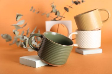 Farklı seramik kupalar, alçı podyumlar ve turuncu arka planda kurumuş okaliptüs dalları.