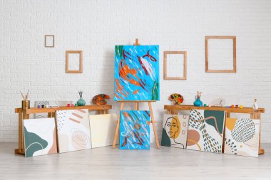 Sanatçı atölyesinin içinde farklı tablolar ve ahşap banklarda sanat malzemeleri var.