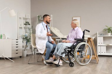 Erkek doktor hastanede tekerlekli sandalyede olgun bir kadınla konuşuyor.