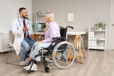 Erkek doktor hastanede tekerlekli sandalyede olgun bir kadınla konuşuyor.