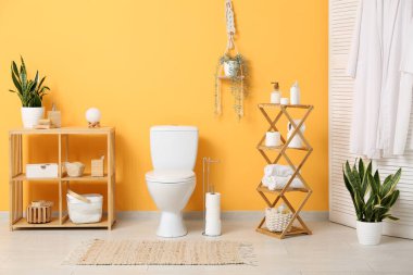 Tuvaletin içinde klozet, raf üniteleri ve sarı duvarın yanında bitkiler var.