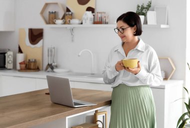 Mutfaktaki masada çay ve dizüstü bilgisayarı olan olgun bir kadın.