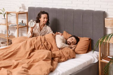 Çalar saati olan genç bir kadın kocasını yatak odasında uyandırıyor.