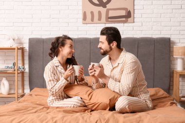 Evdeki yatakta pijamalı, kahveli mutlu bir çift oturuyor.