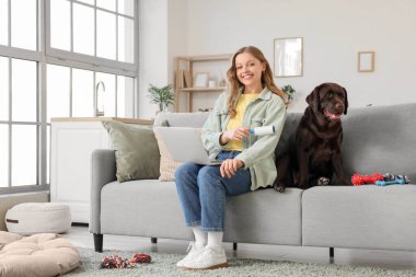 Tişörtünü pamuklu silindirle temizleyen genç bir kadın ve evdeki koltukta Labrador köpeği.