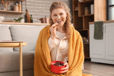 Güzel, genç bir kadın, rahat bir battaniye ve bir kutu çikolata şekeriyle evde oturuyor.