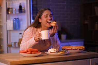 Geceleri mutfaktaki masada kek ve farklı yiyeceklerle mutlu genç bir kadın.