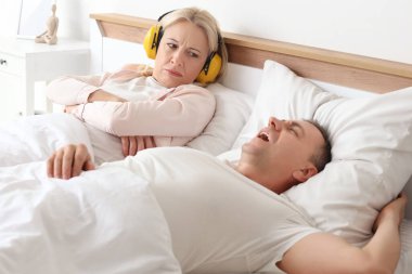 Duyma koruyucuları içinde sinirli olgun bir kadın horlayan kocasıyla evde yatakta yatıyor.