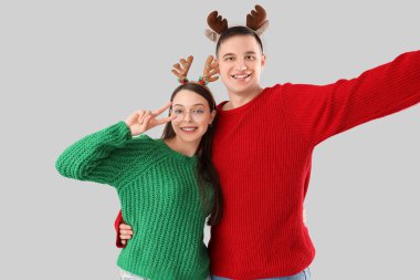 Gri arka planda selfie çeken kalın kazaklı ve Noel bantlı sevimli genç bir çift.