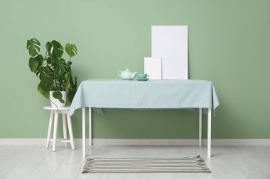 Yeşil duvarın yanındaki masa örtüsünün üzerinde boş çerçeveli çaydanlık.