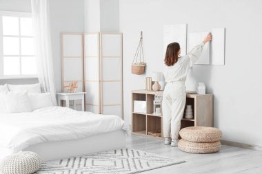 Young woman hanging painting on light wall in bedroom, back view