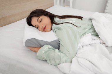 Pijamalı genç bir kadın evdeki rahat yatakta yumuşak ortopedik yastığa uzanıyor.