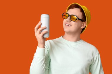 Güneş gözlüklü genç adam, turuncu arka planda bir kutu gazoz.