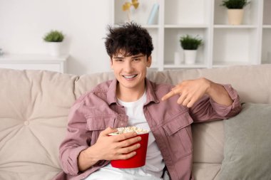 Genç adam film izliyor ve oturma odasındaki kanepede patlamış mısırı işaret ediyor.