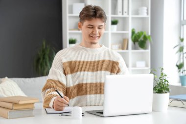 Bilgisayarlı erkek öğrenci evde ders çalışırken not alıyor.