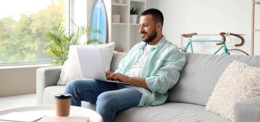 Genç adam evinde dizüstü bilgisayarla kanepede oturuyor.